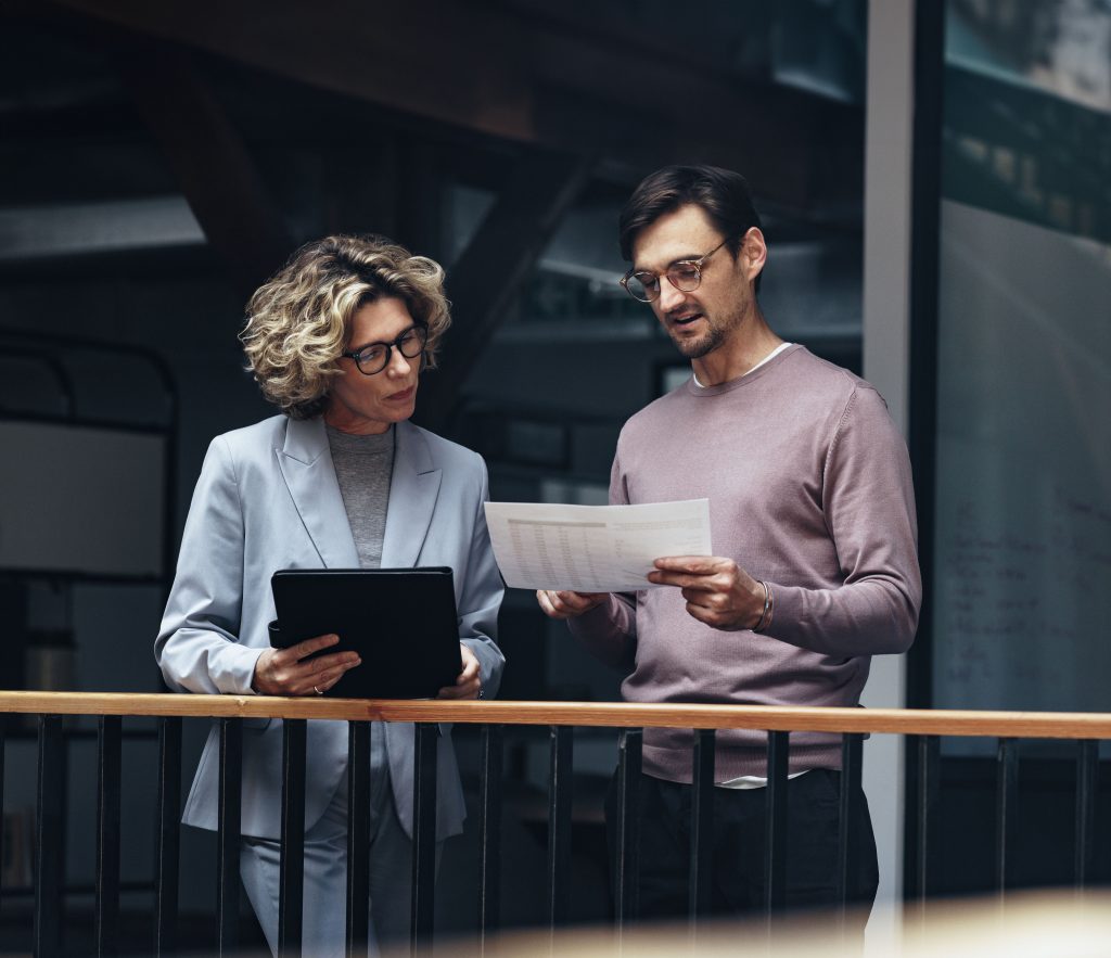 Business people reading a document together. Two professionals analyse a financial report in an office. Business colleagues discuss with each other while standing on an interior balcony.